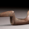 Three-quarter view of a birdstone made of smoothed brown stone with grey marbling that could fit in the hand. There is a long pointed beak, slim body and upright tail.