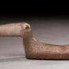 Side view of a birdstone made of smoothed brown stone with grey marbling that could fit in the hand. There is a long pointed beak, slim body and upright tail.